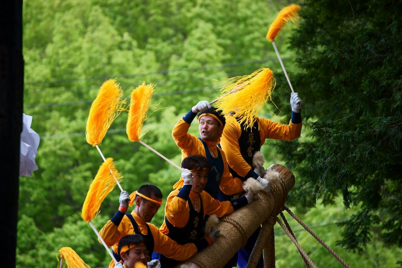 Peserta festival mengenakan kostum oranye menari di atas perahu dengan tongkat bulu kuning di latar belakang gunung hijau
