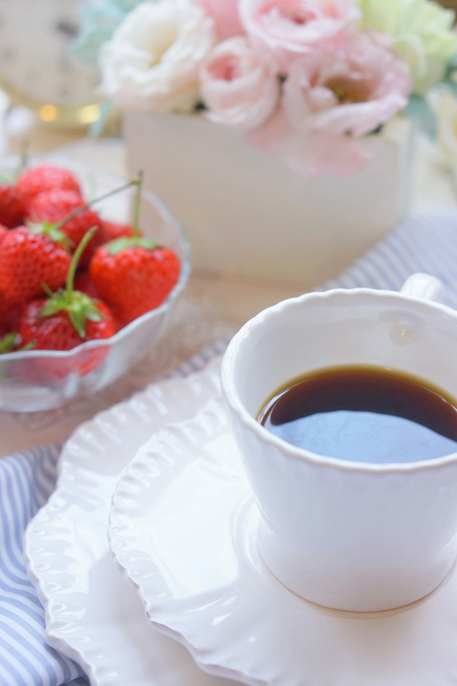 กาแฟในถ้วยสีขาวพร้อมกับสตรอเบอรี่สดบนจานและการจัดดอกไม้ในพื้นหลัง