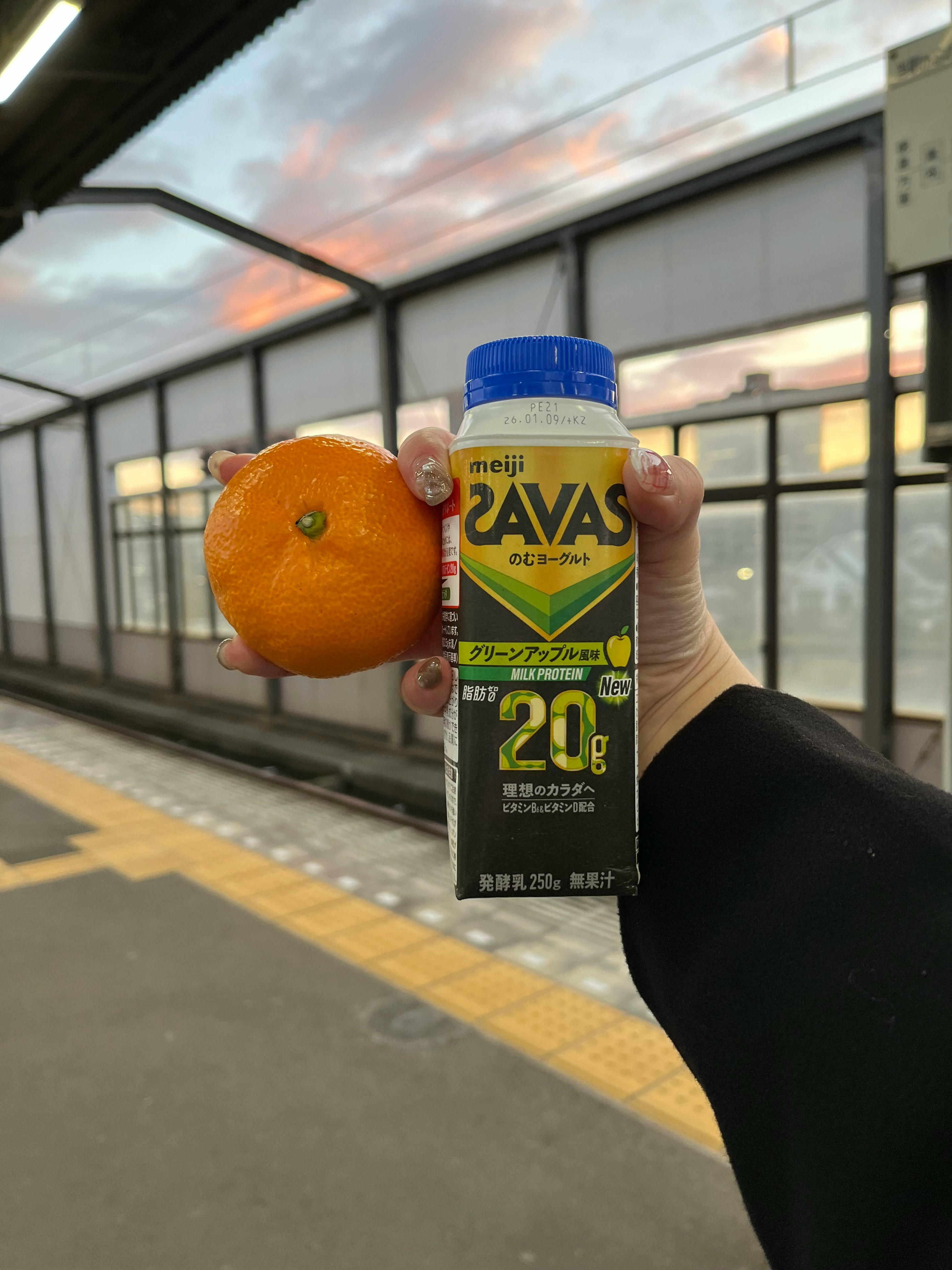 駅のホームで手に持つ飲料パックとオレンジのキーホルダーのある風景