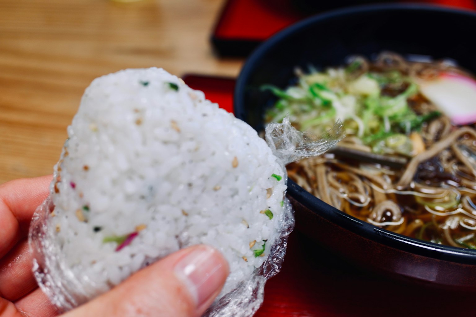 Une main tenant un onigiri avec un bol de ramen en arrière-plan