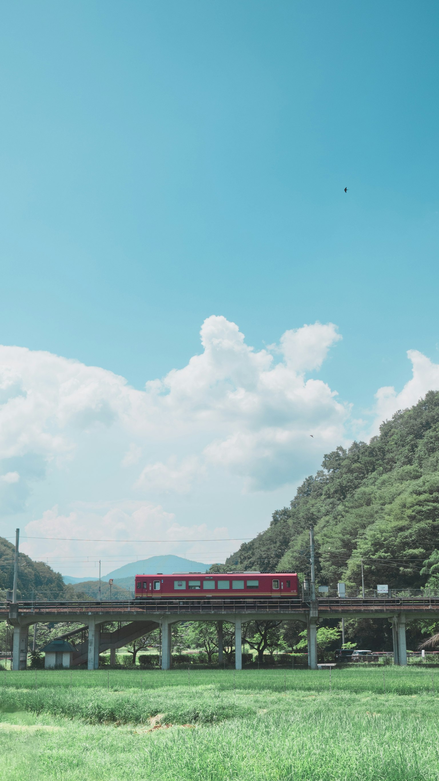 Kereta merah melintasi jembatan di bawah langit biru dengan pemandangan hijau