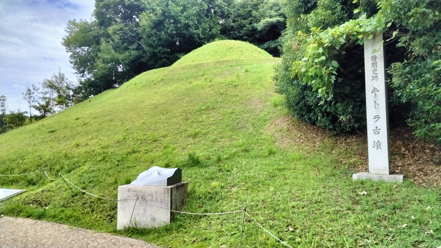 Sebuah gundukan kecil yang ditumbuhi rumput dengan tanda batu di dekatnya dalam lanskap hijau