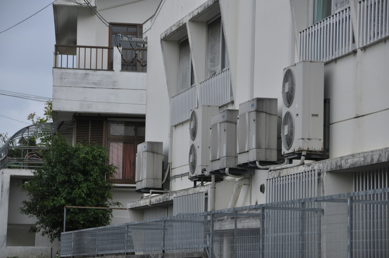 多台空调装置安装在白色建筑物的外墙上，旁边有围栏