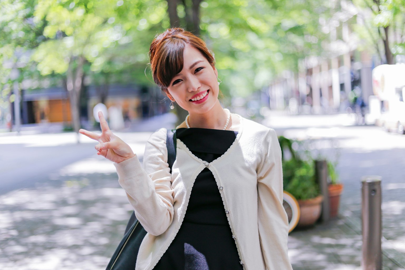 หญิงสาวยิ้มทำสัญลักษณ์แห่งสันติในสวนสาธารณะมีต้นไม้สีเขียวและถนนสว่างในพื้นหลัง