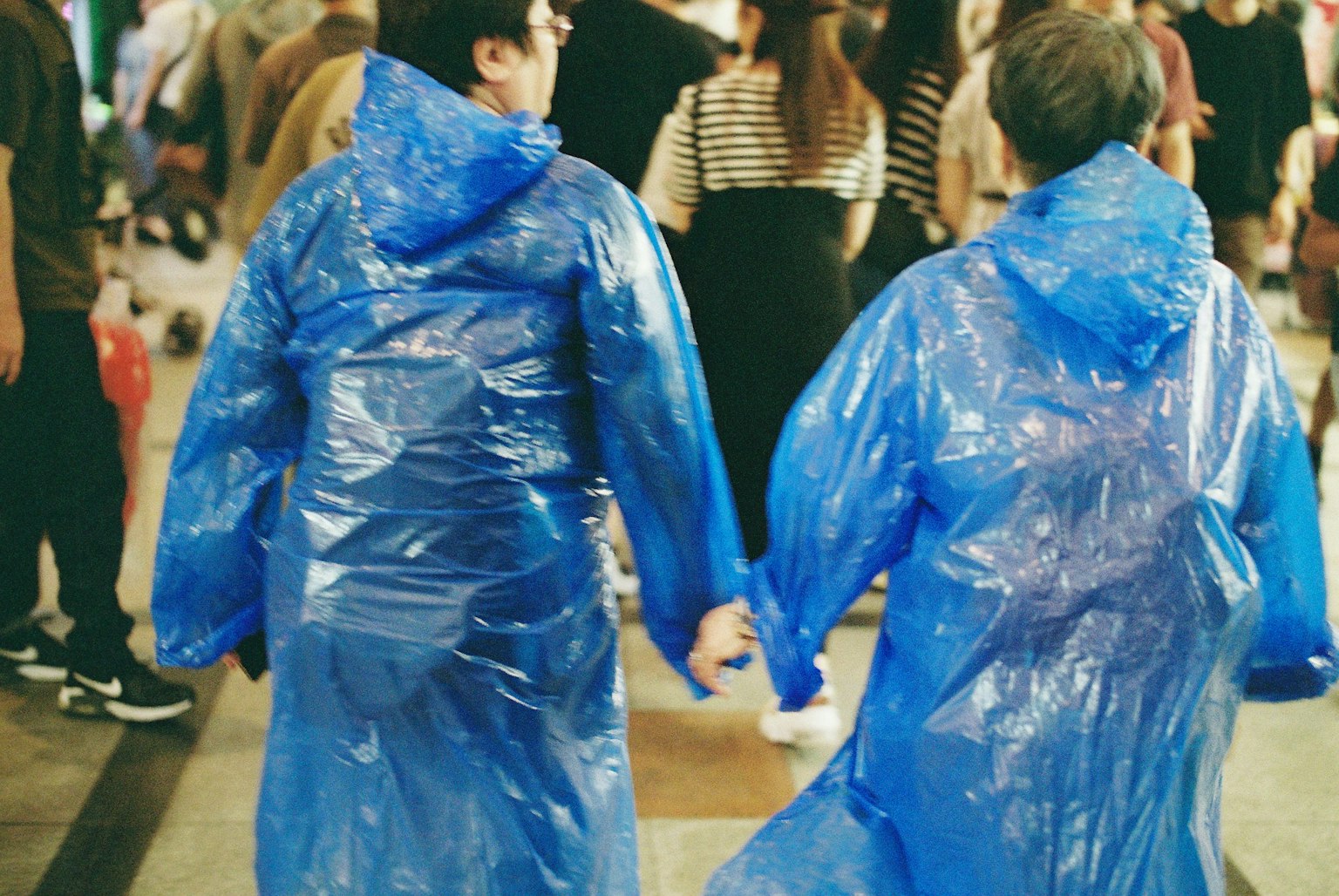 Zwei Personen in blauen Regenmänteln, die Händchen haltend mit einer Menschenmenge im Hintergrund