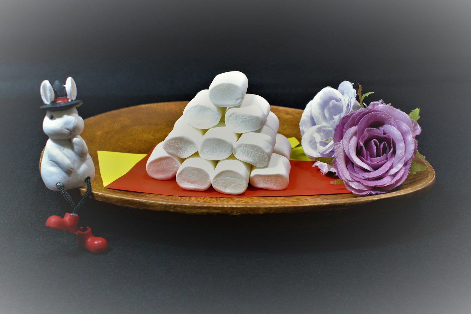 A wooden plate featuring a pyramid of white marshmallows alongside a purple rose and a cute rabbit figurine