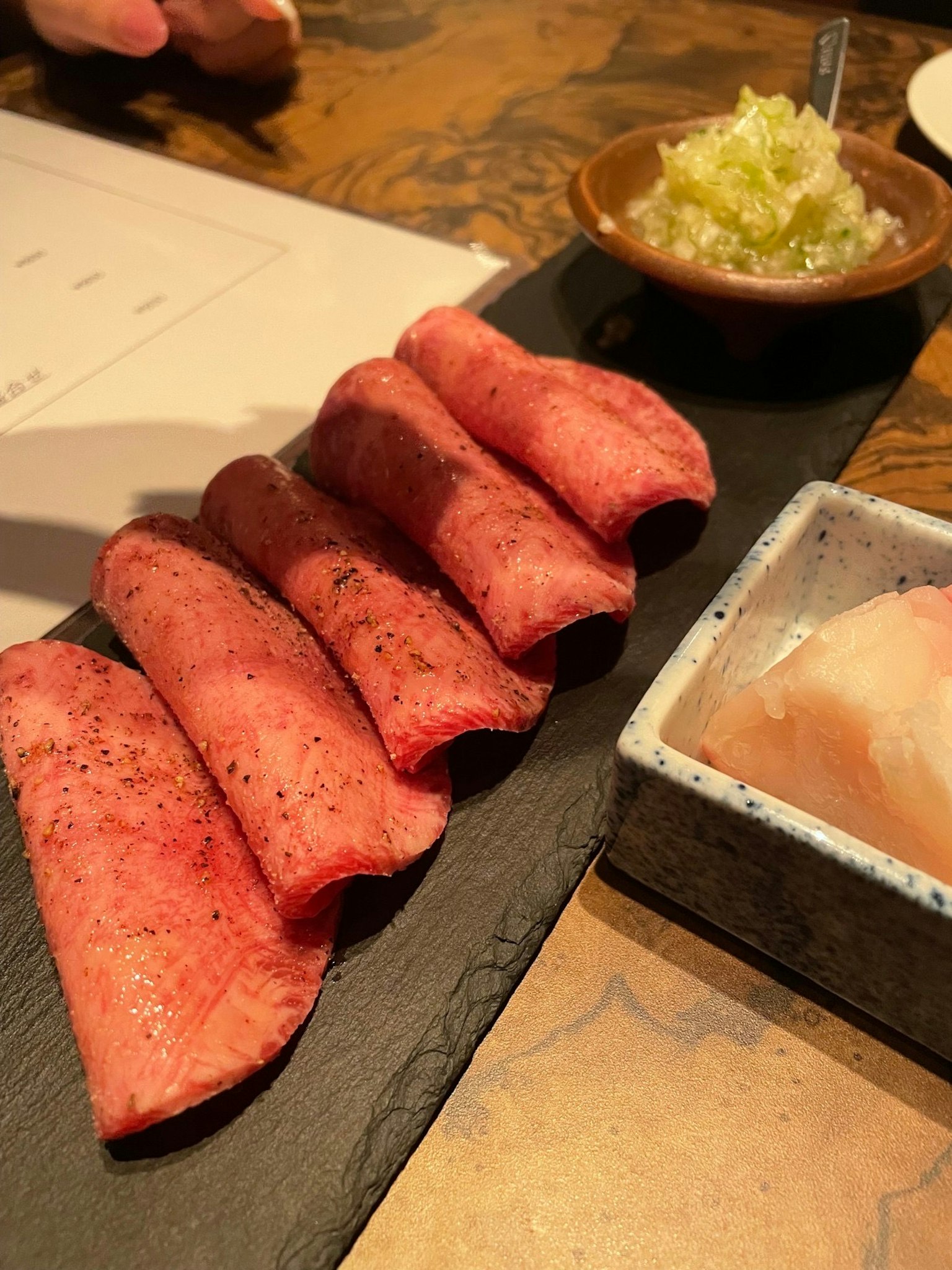 五片大理石紋肉在黑色盤子上，旁邊有一個小碗的配料