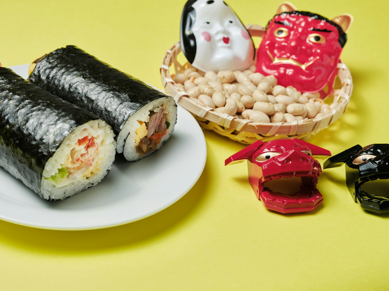 Gambar menampilkan sushi gulung di piring dengan figur hias dan kacang tanah di latar belakang kuning