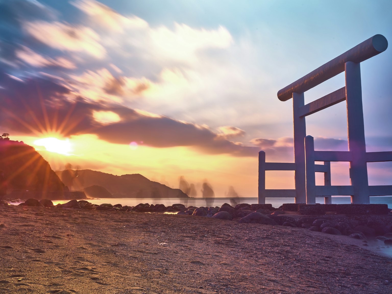Weißes Torii auf einem Strand bei Sonnenuntergang mit Bergen im Hintergrund