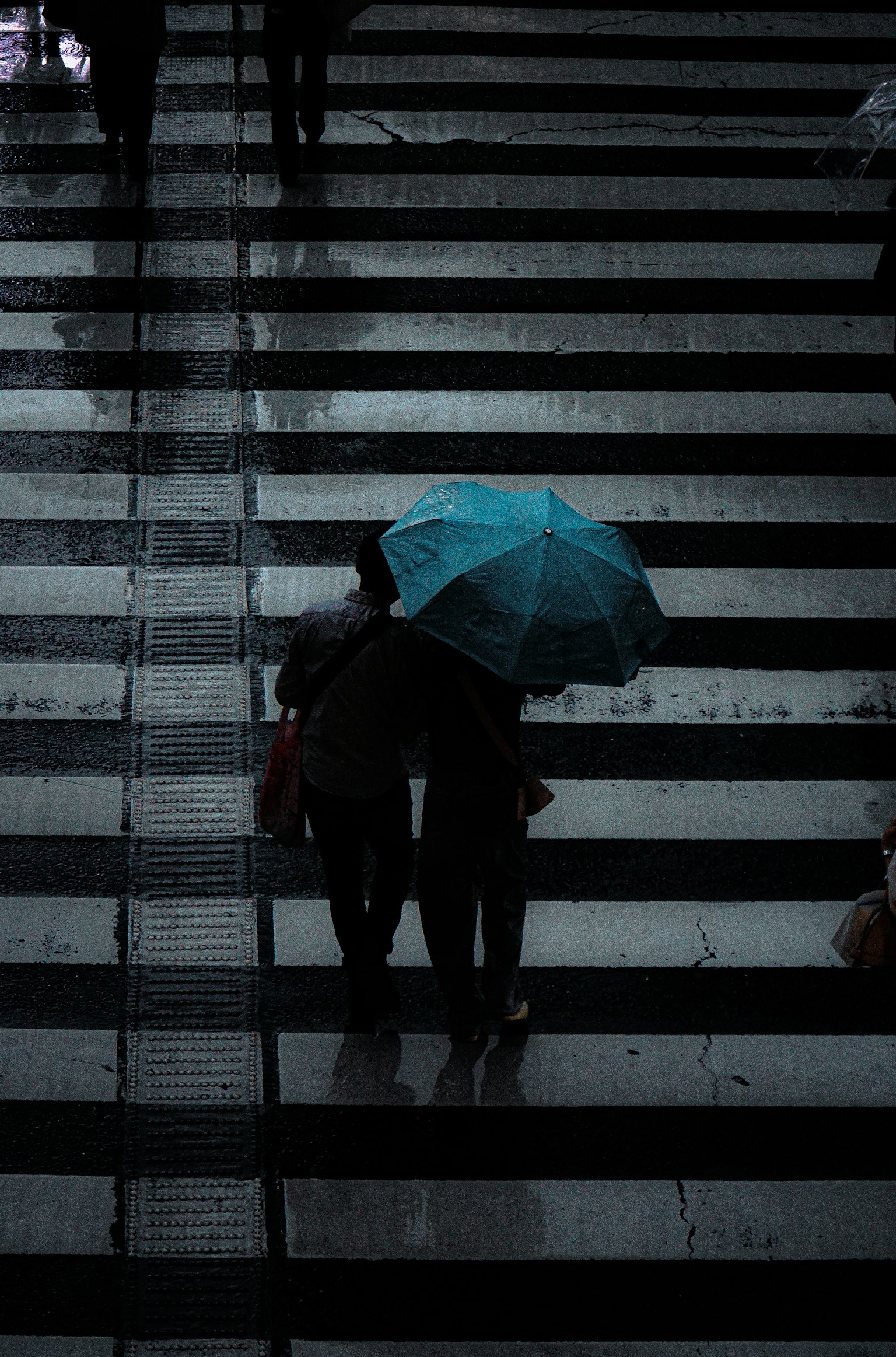 Silhouette of two people holding a turquoise umbrella walking on a wet crosswalk