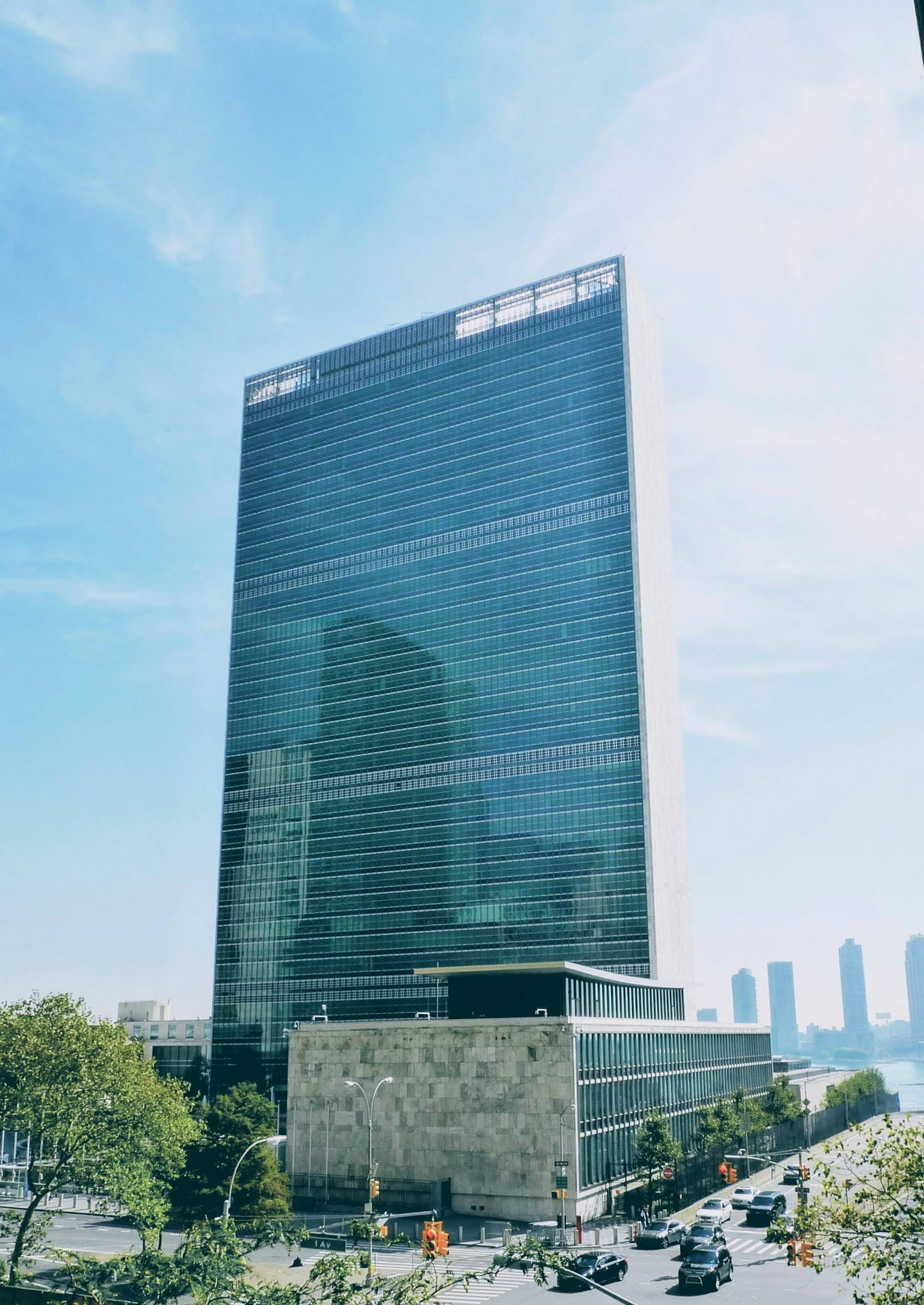 Modern skyscraper with glass facade under blue sky and clouds, surrounded by greenery