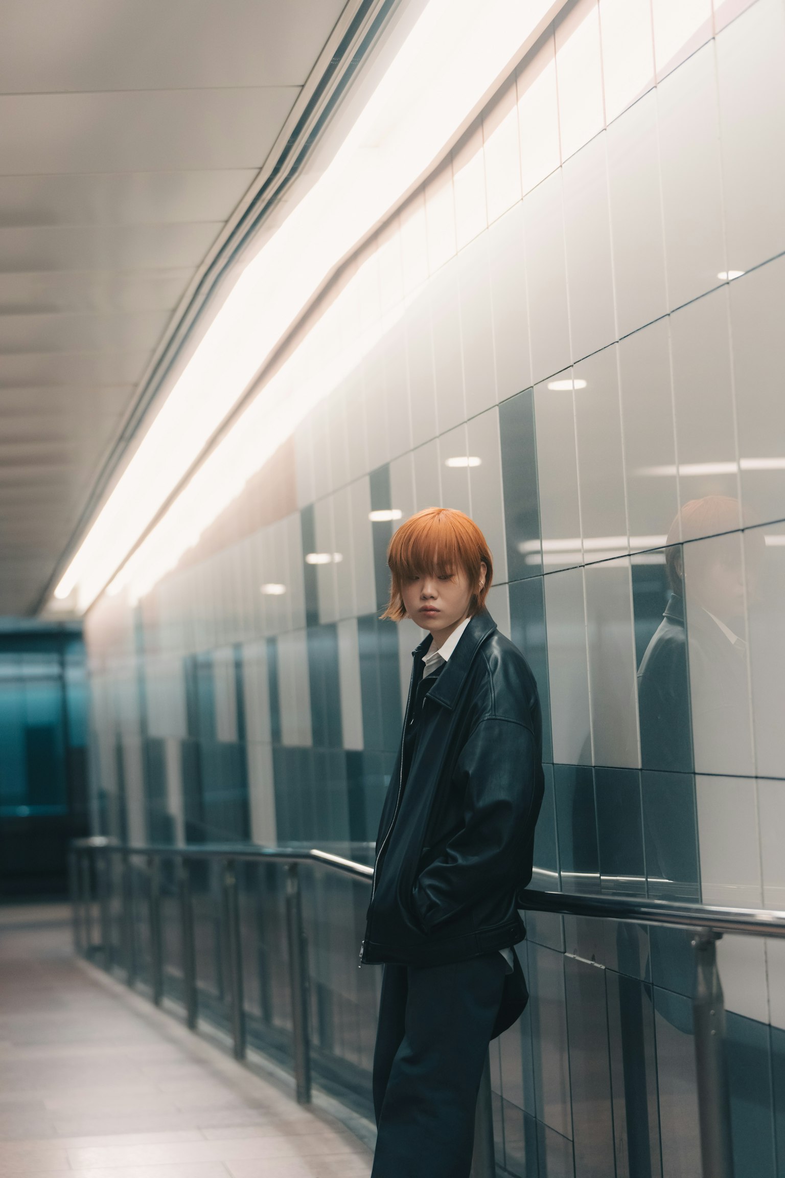 Un joven con cabello naranja de pie en un pasaje subterráneo con una chaqueta negra fondo de azulejos modernos