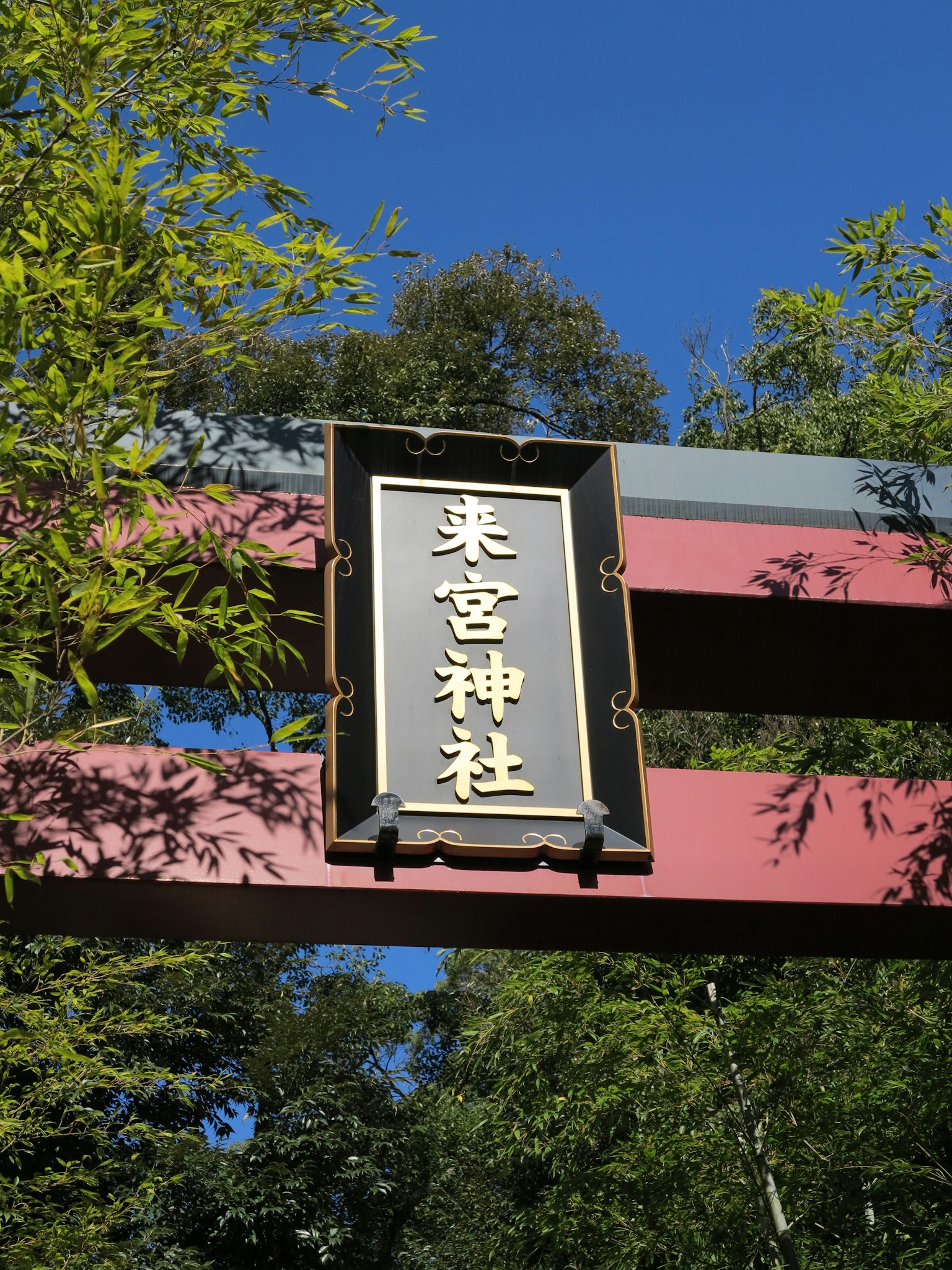 Sign of a shrine above a red torii gate with blue sky and green foliage