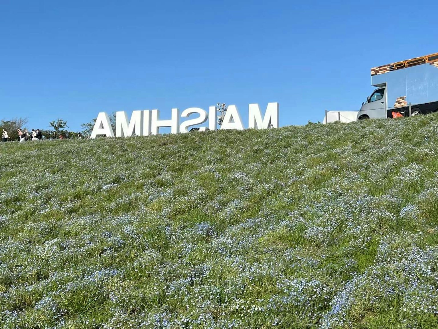 ป้ายขนาดใหญ่สีขาวเขียนว่า 'MAISHIMA' บนหญ้าสีเขียวใต้ท้องฟ้าสีฟ้า