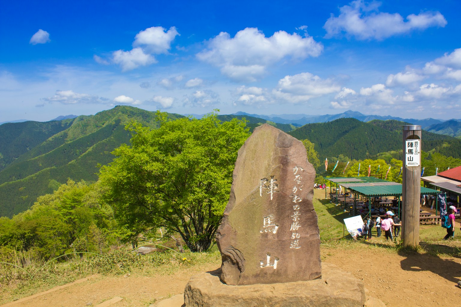 Scenic view from a mountain top featuring a large stone monument with inscriptions surrounded by lush green mountains and visitors