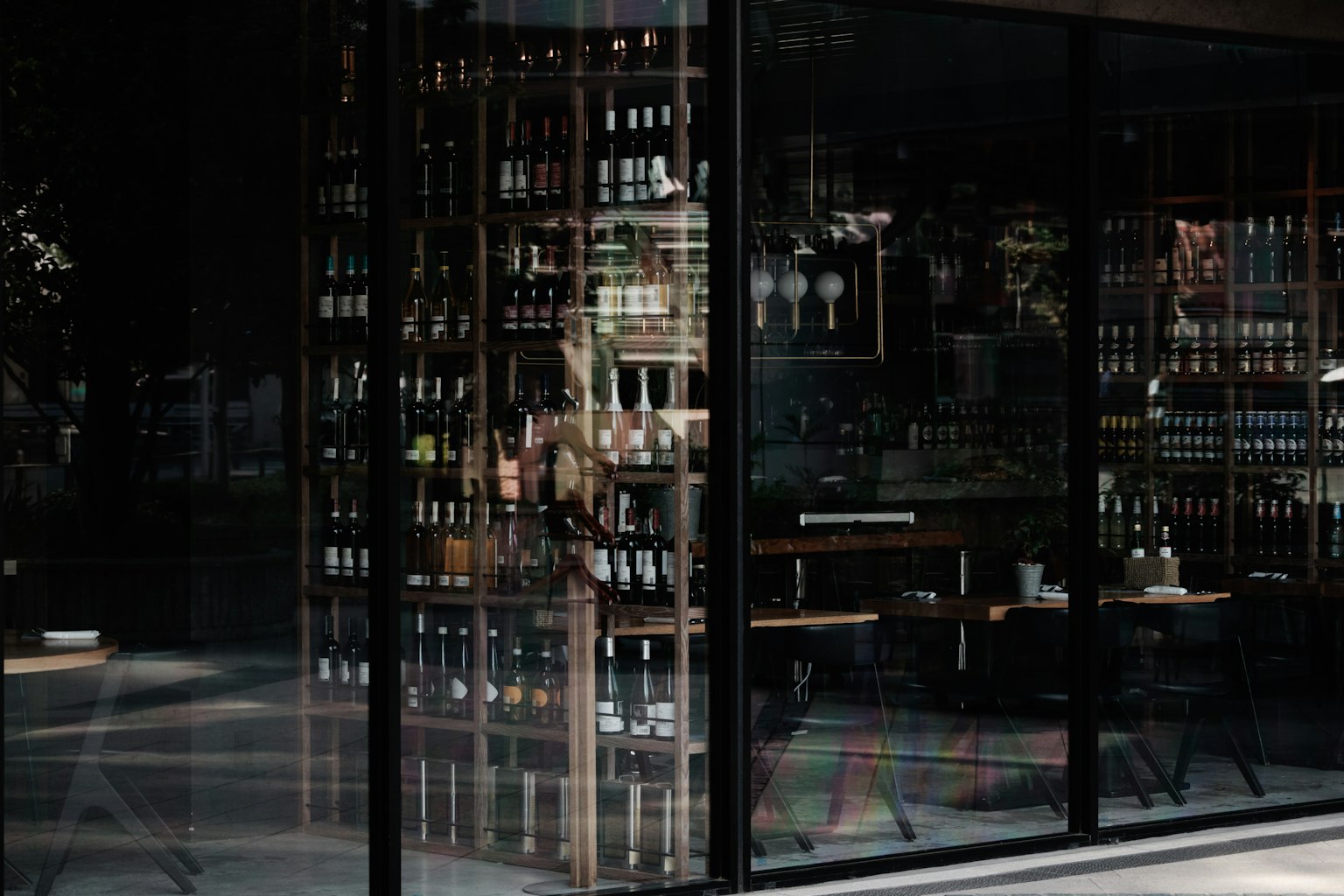 Vue intérieure d'un restaurant à travers une vitre montrant des étagères remplies de bouteilles et des tables disposées
