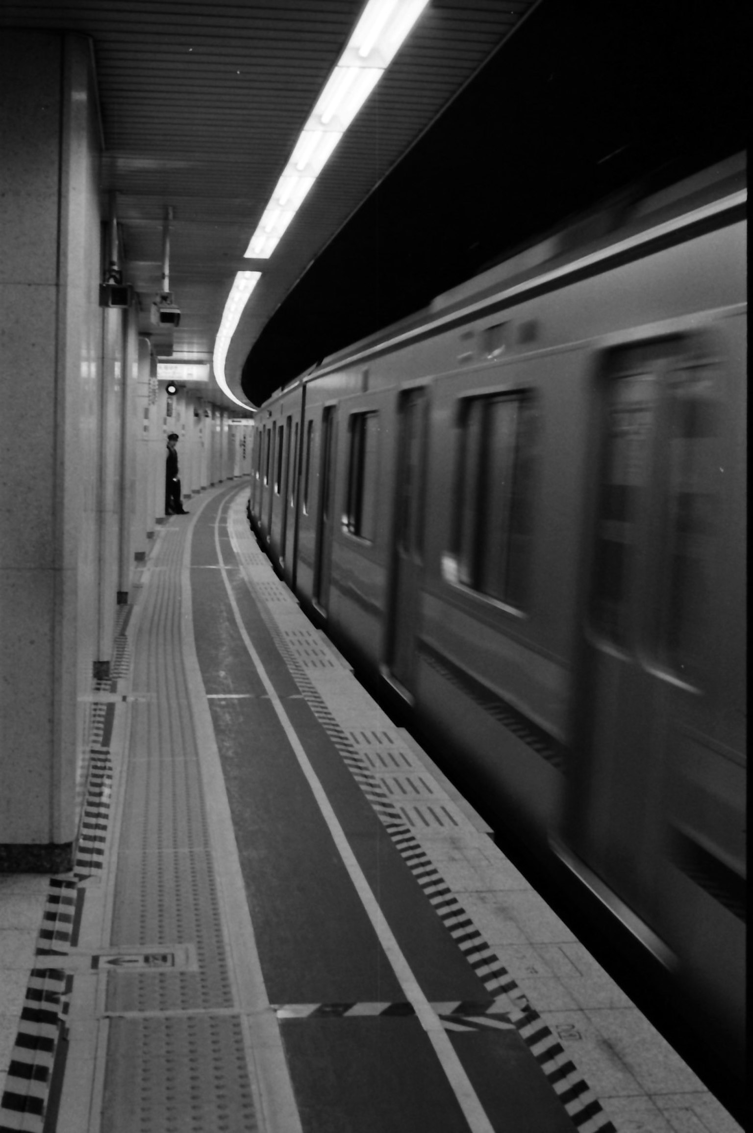 Stasiun kereta bawah tanah hitam putih dengan platform melengkung dan kereta yang lewat