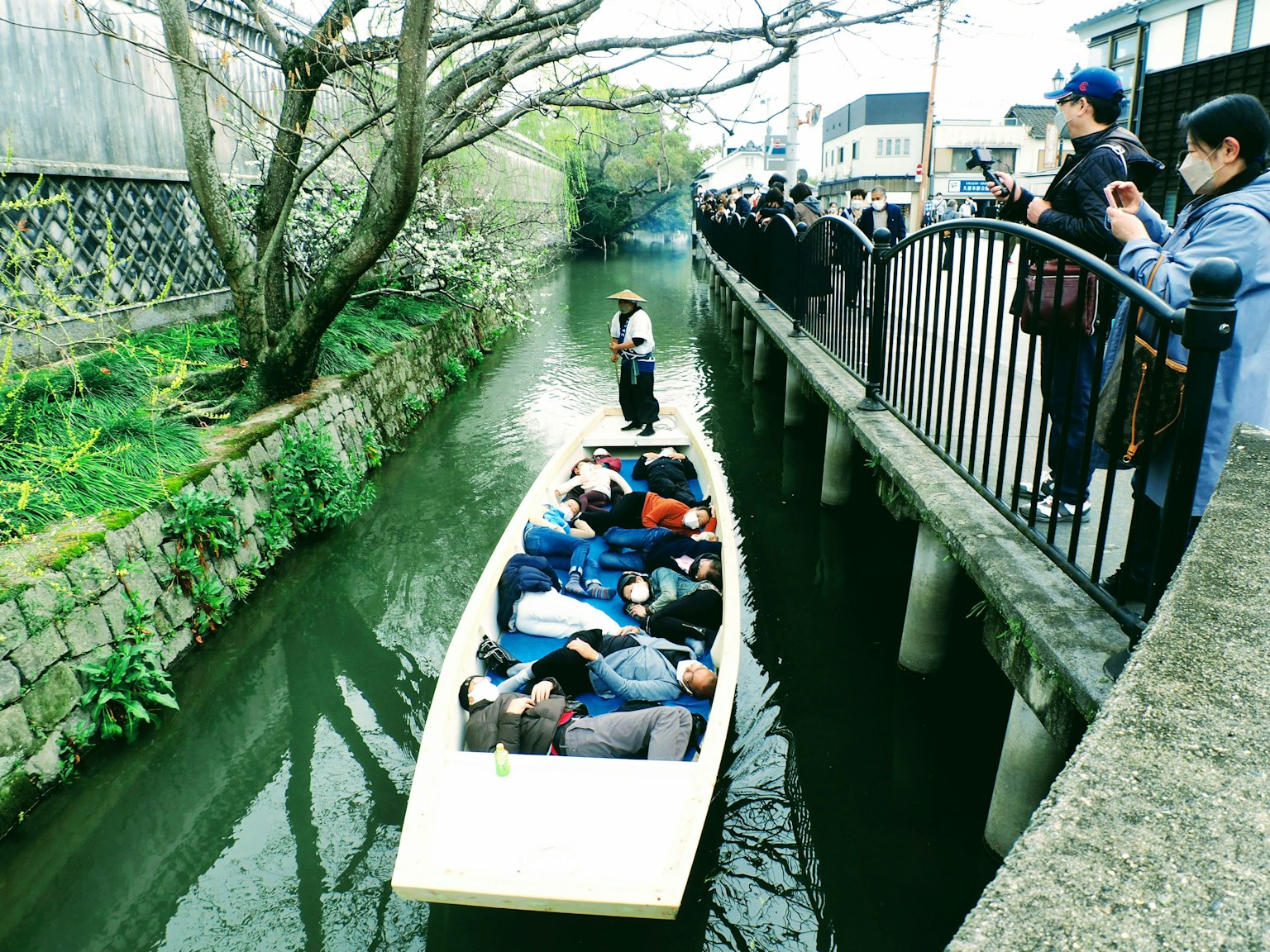 ภาพเรือในแม่น้ำพร้อมนักท่องเที่ยวที่อยู่ริมฝั่ง คนที่นอนอยู่ในเรือ ต้นซากุระอยู่ด้านหลัง