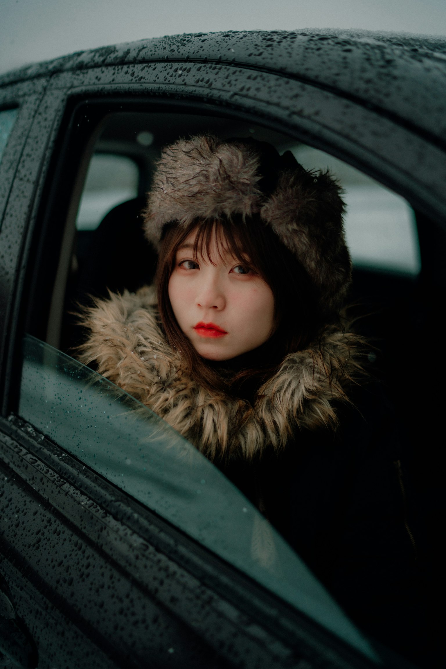 Mujer vista a través de una ventana de coche usando un abrigo cálido y un sombrero en un entorno nevado con labios rojos