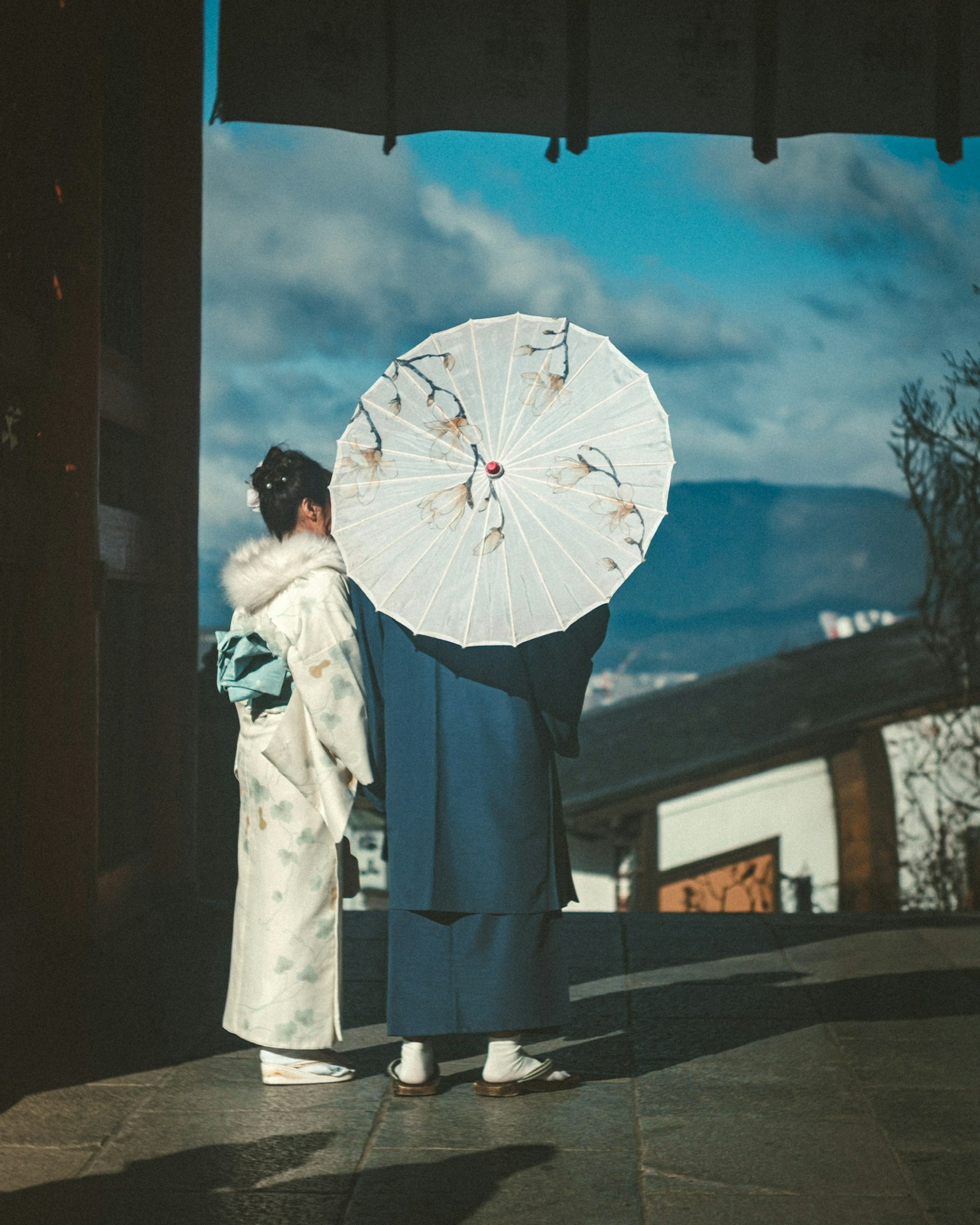 Two individuals in traditional clothing holding a white umbrella against a scenic backdrop