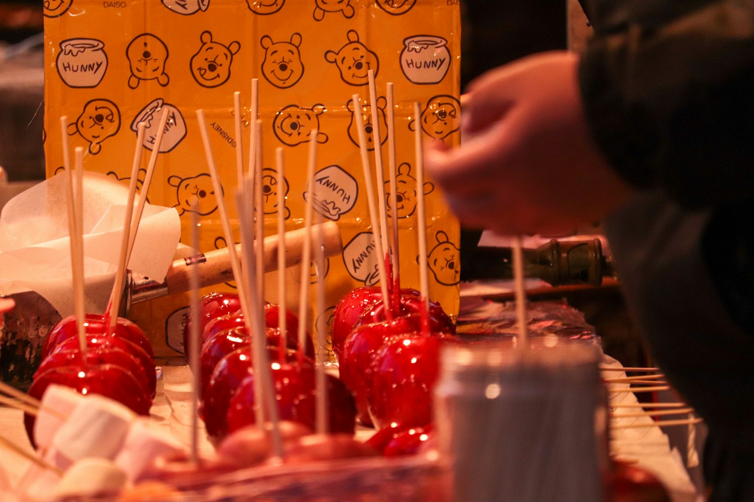 Close-up of candied apples on sticks at a market stall with a hand reaching for them and a colorful illustrated box in the background