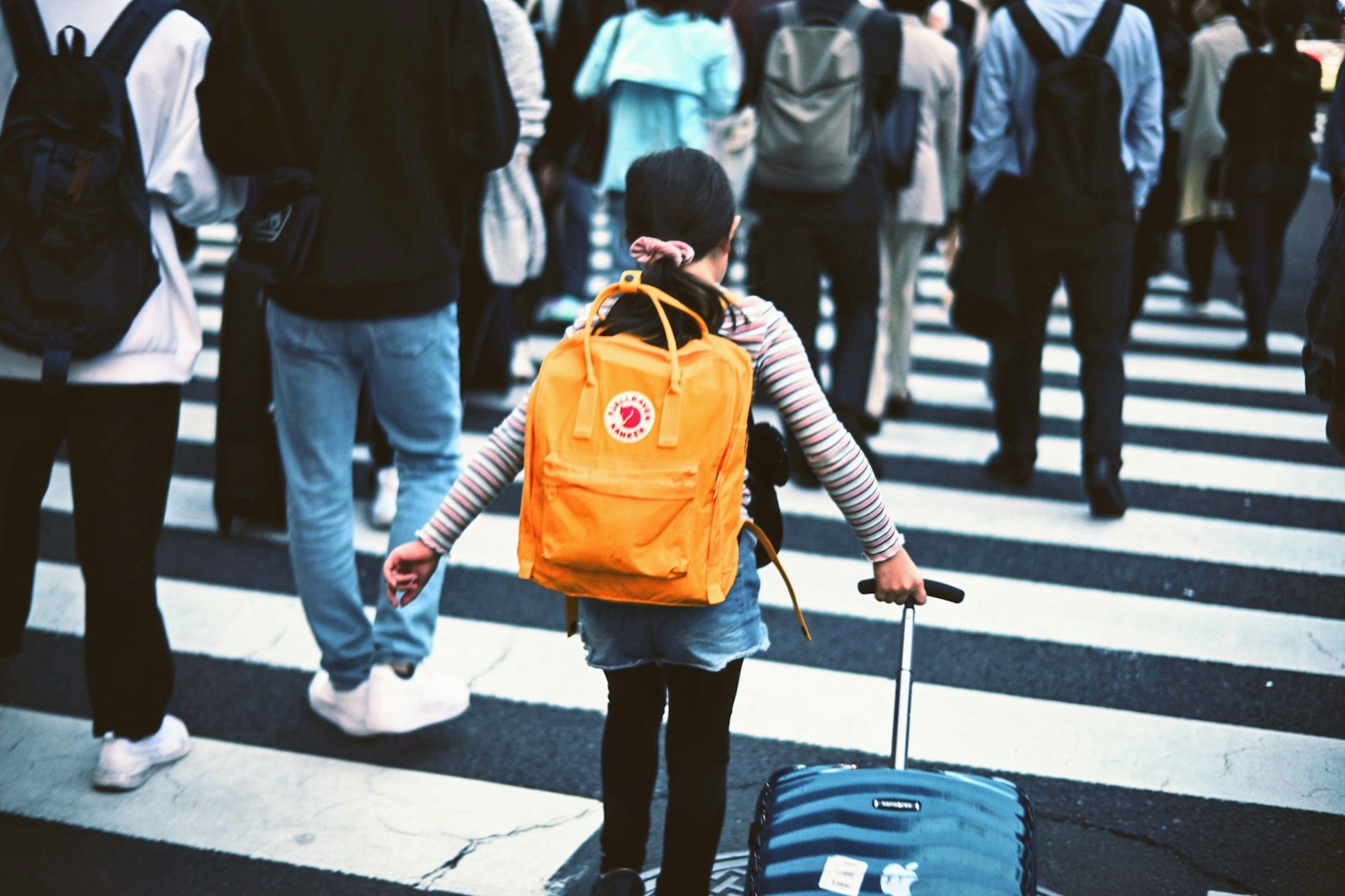 A child crossing the street with a suitcase pulling an orange backpack