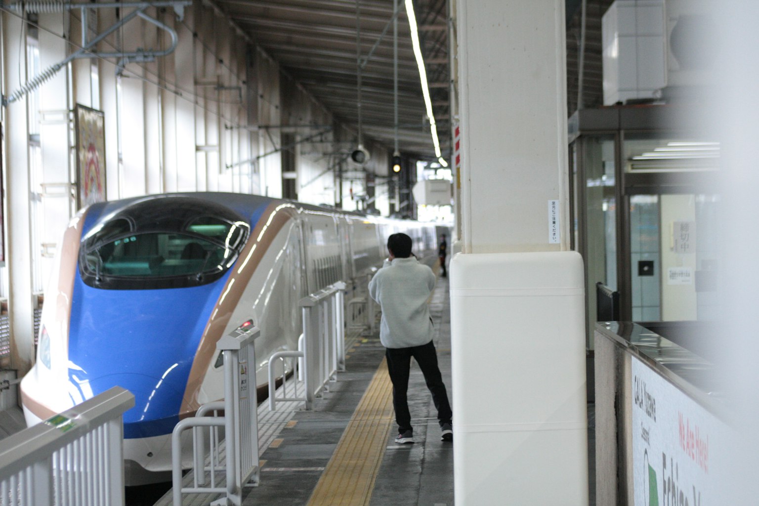 ชายคนหนึ่งยืนอยู่บนชานชาลาของสถานีที่มีรถไฟสีน้ำเงินจอดอยู่