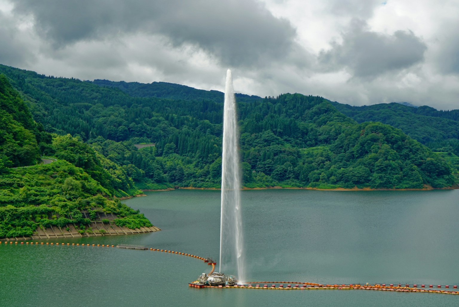 Une fontaine projetant de l'eau dans les airs au centre d'un lac entouré de montagnes