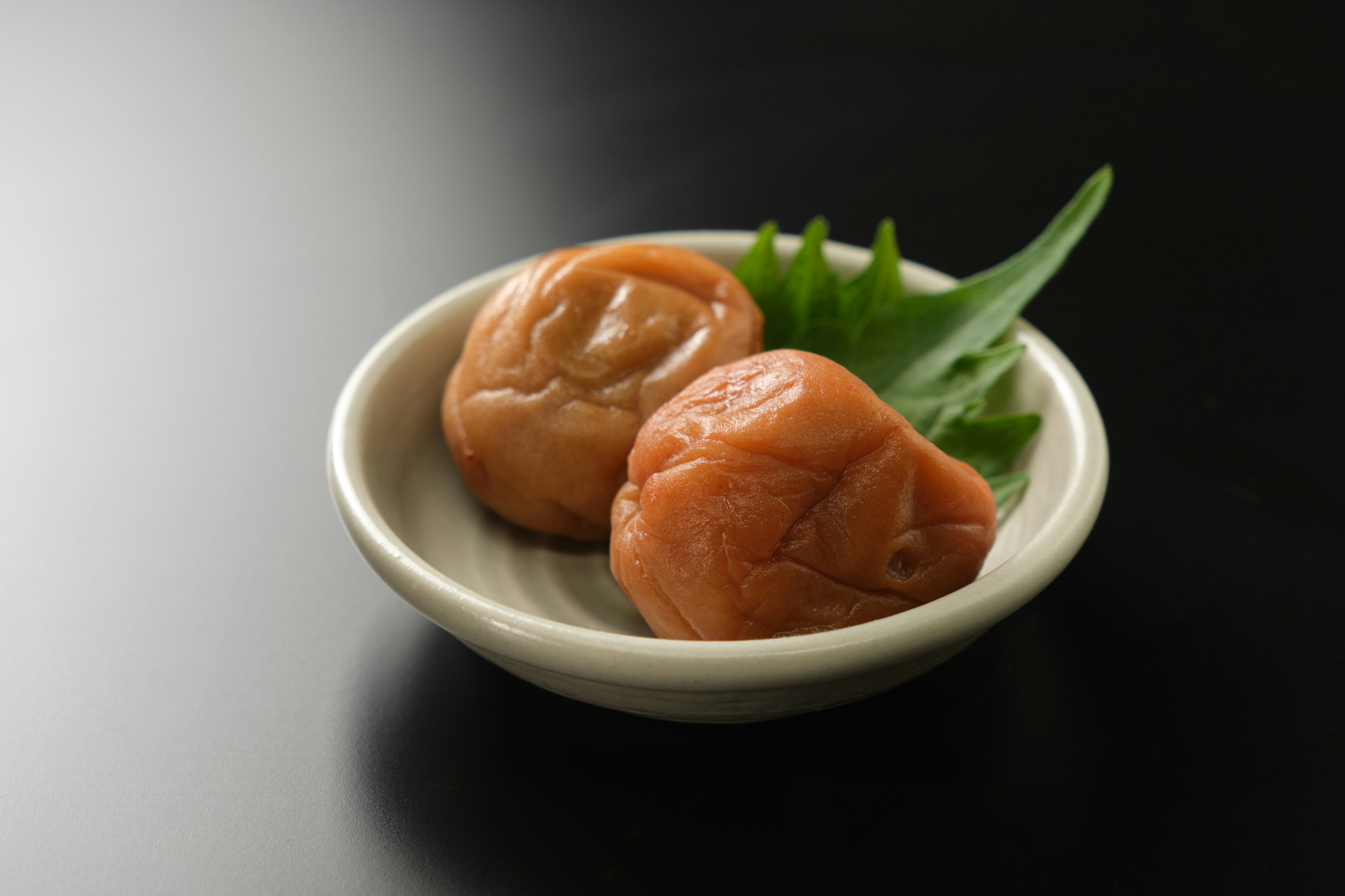 Dua umeboshi di atas piring putih kecil dengan daun hijau