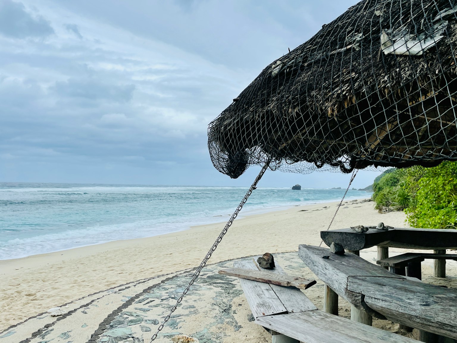 Pemandangan pantai dengan meja kayu dan tempat berlindung tertutup jaring yang menghadap laut