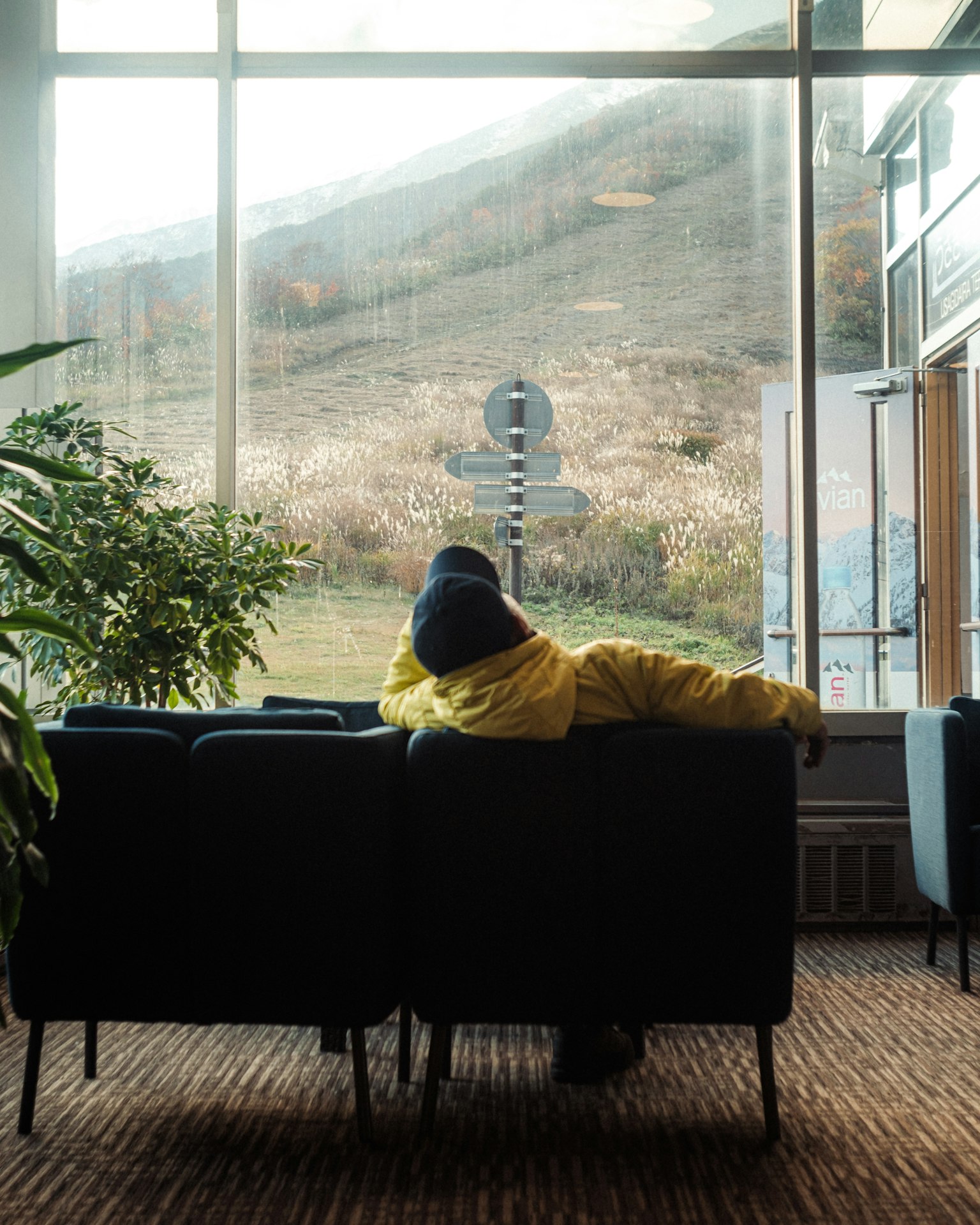 Une personne en veste jaune assise sur un canapé regardant par la fenêtre avec une vue sur la montagne