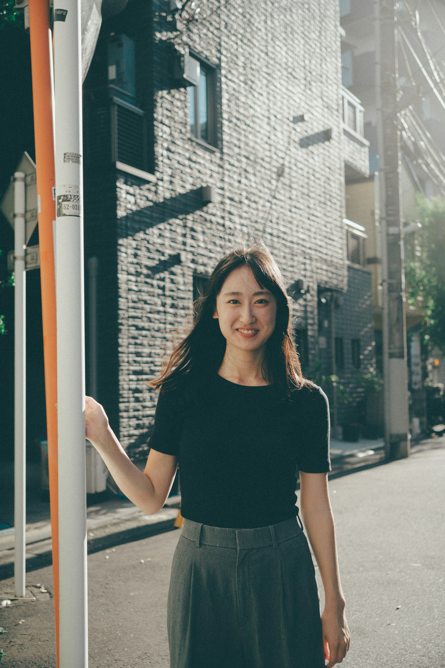 A woman smiling in the street wearing a black t-shirt and gray pants with a brick building in the background