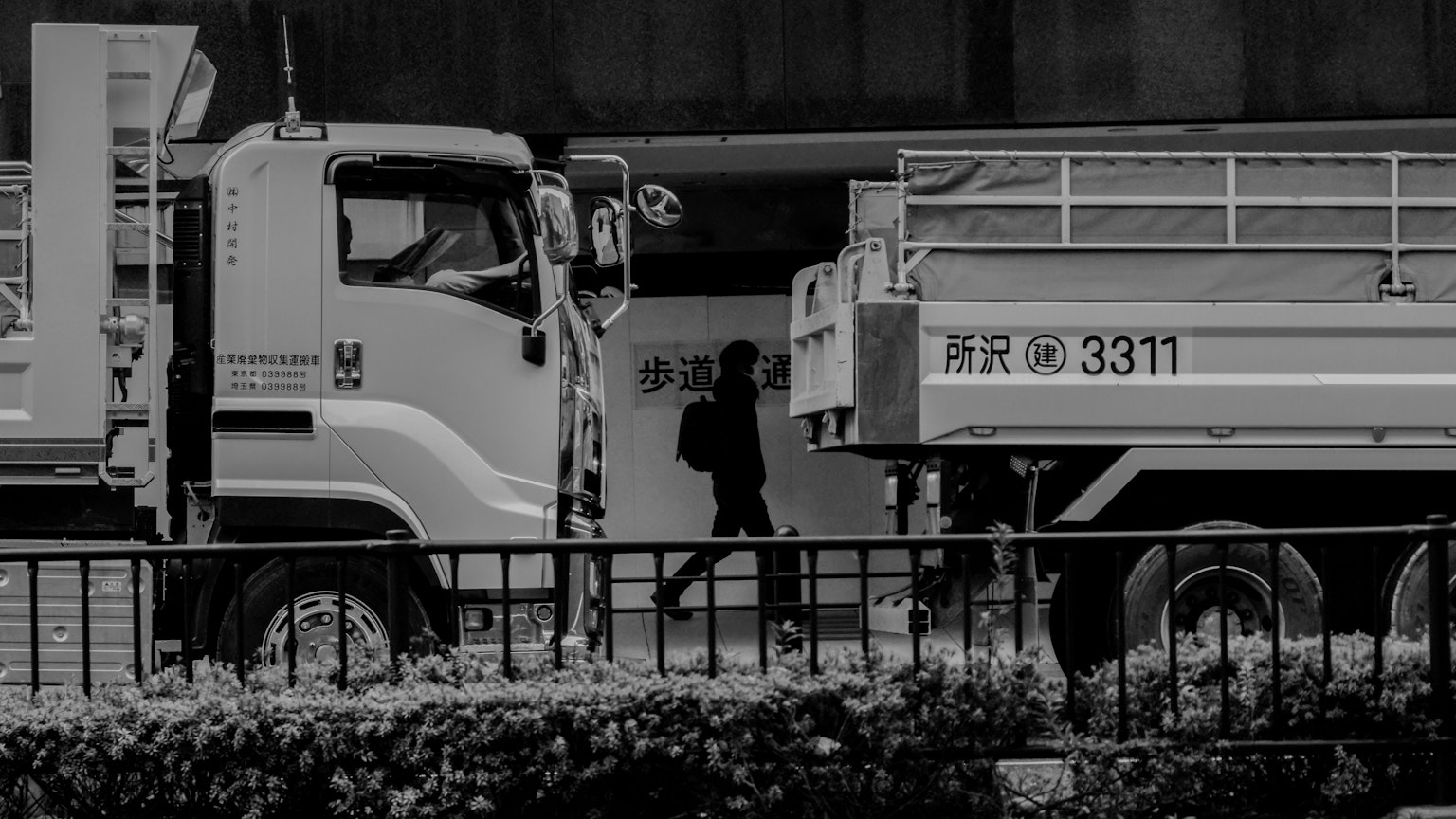 Silueta de una persona caminando junto a camiones en un paisaje urbano en blanco y negro