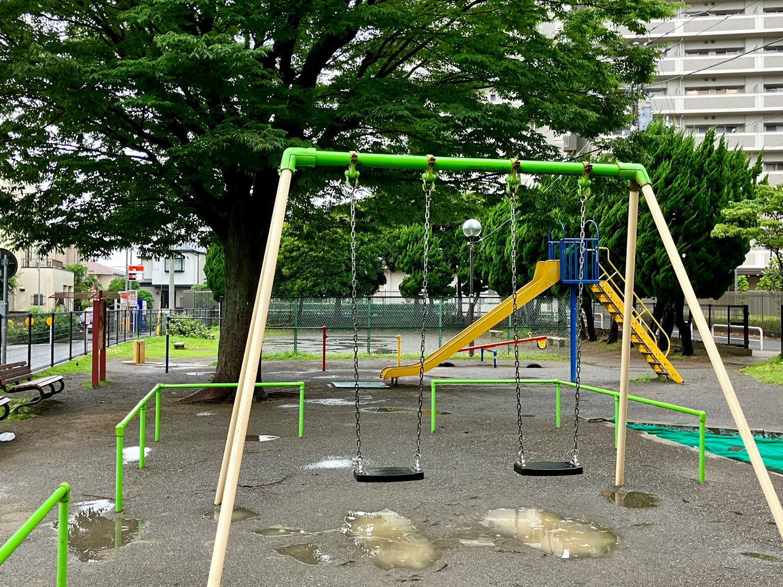 Parque infantil con columpios y tobogán con estructura verde y gran árbol