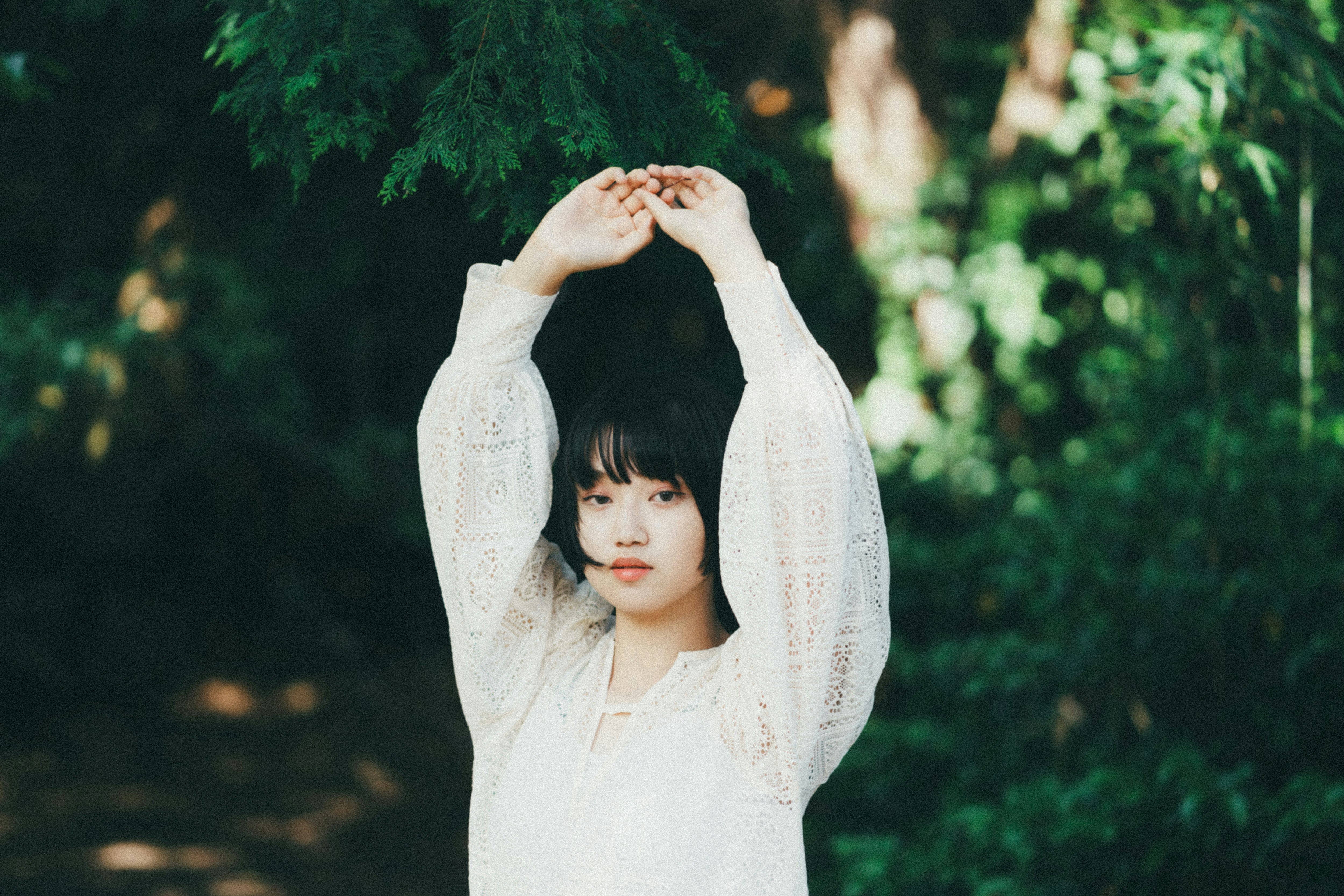 木々の中で両手を頭上に掲げる白い服の女性