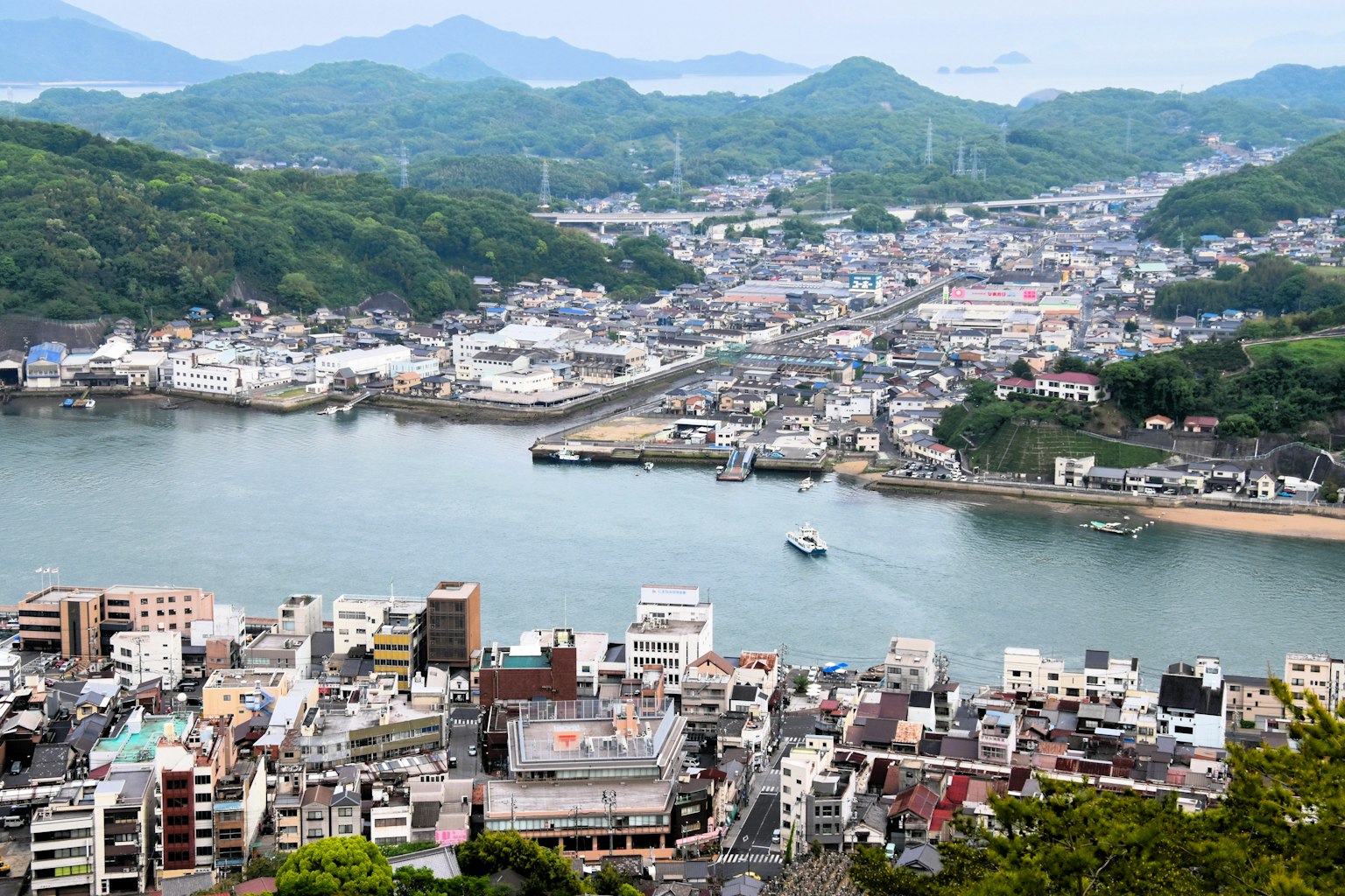 A panoramic view of a coastal town from a mountain featuring green hills and a bay