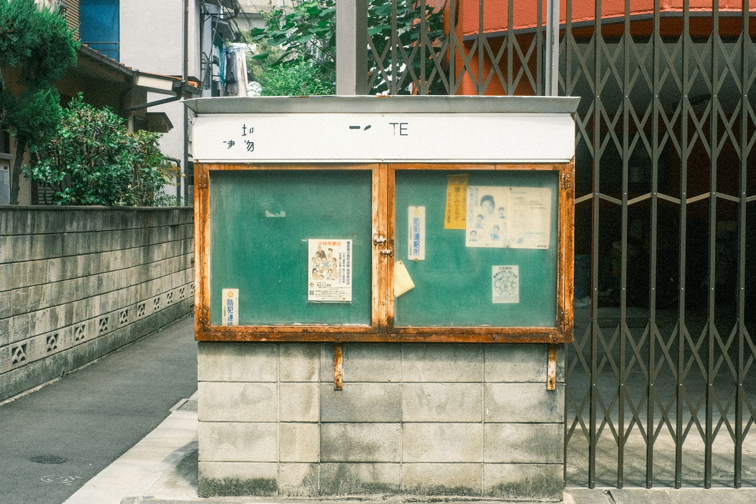 Un vecchio pannello informativo all'angolo di una strada con un muro verde e manifesti scollati