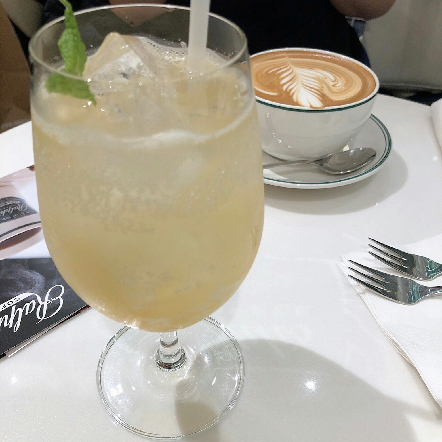 Một ly nước uống màu vàng nhạt có đá và một tách cà phê latte nghệ thuật ở phía sau trong quán cà phê