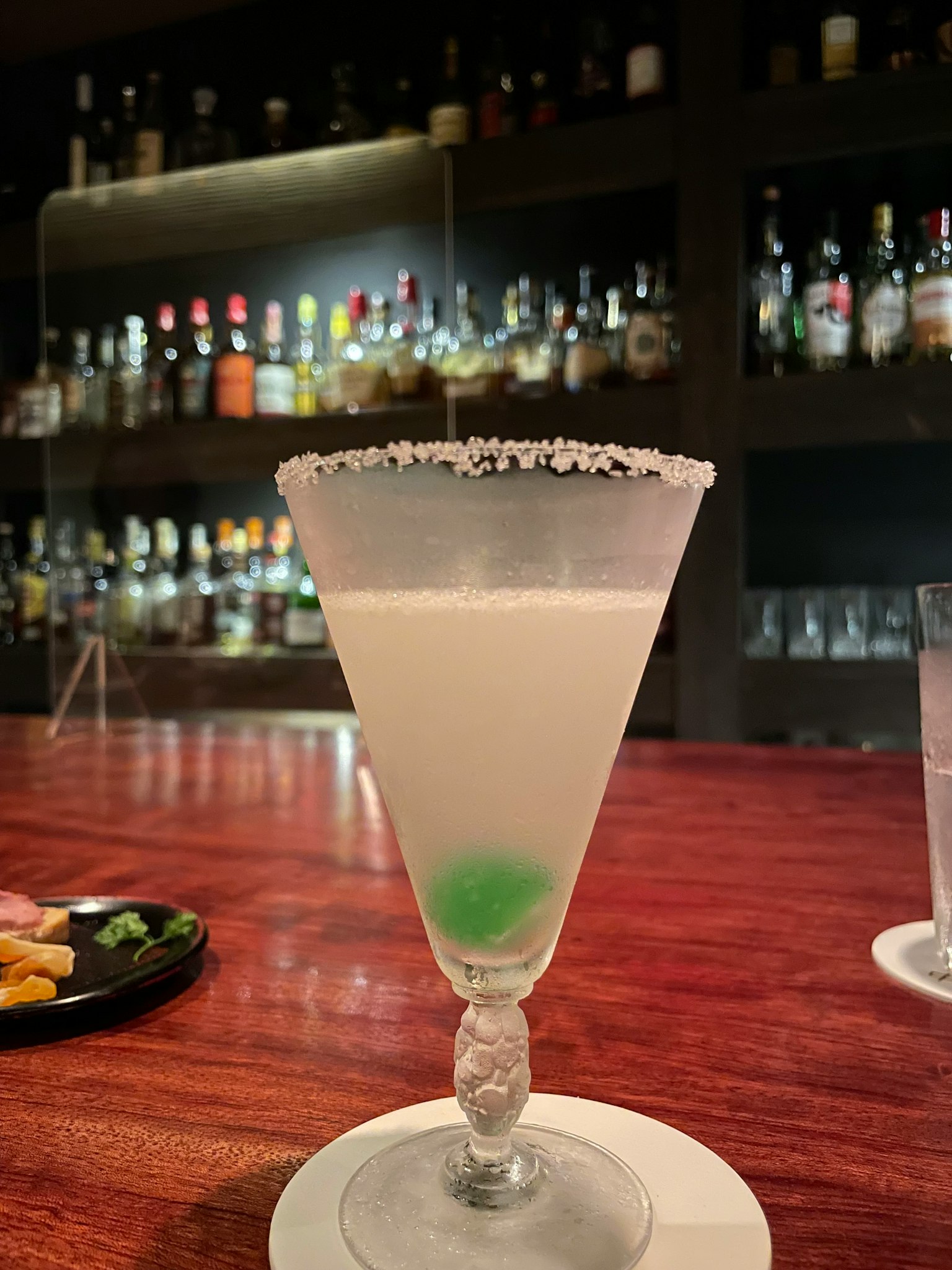 Bicchiere da cocktail con una bevanda bianca e una guarnizione di caramella verde, sfondo con varie bottiglie di liquore in un bar