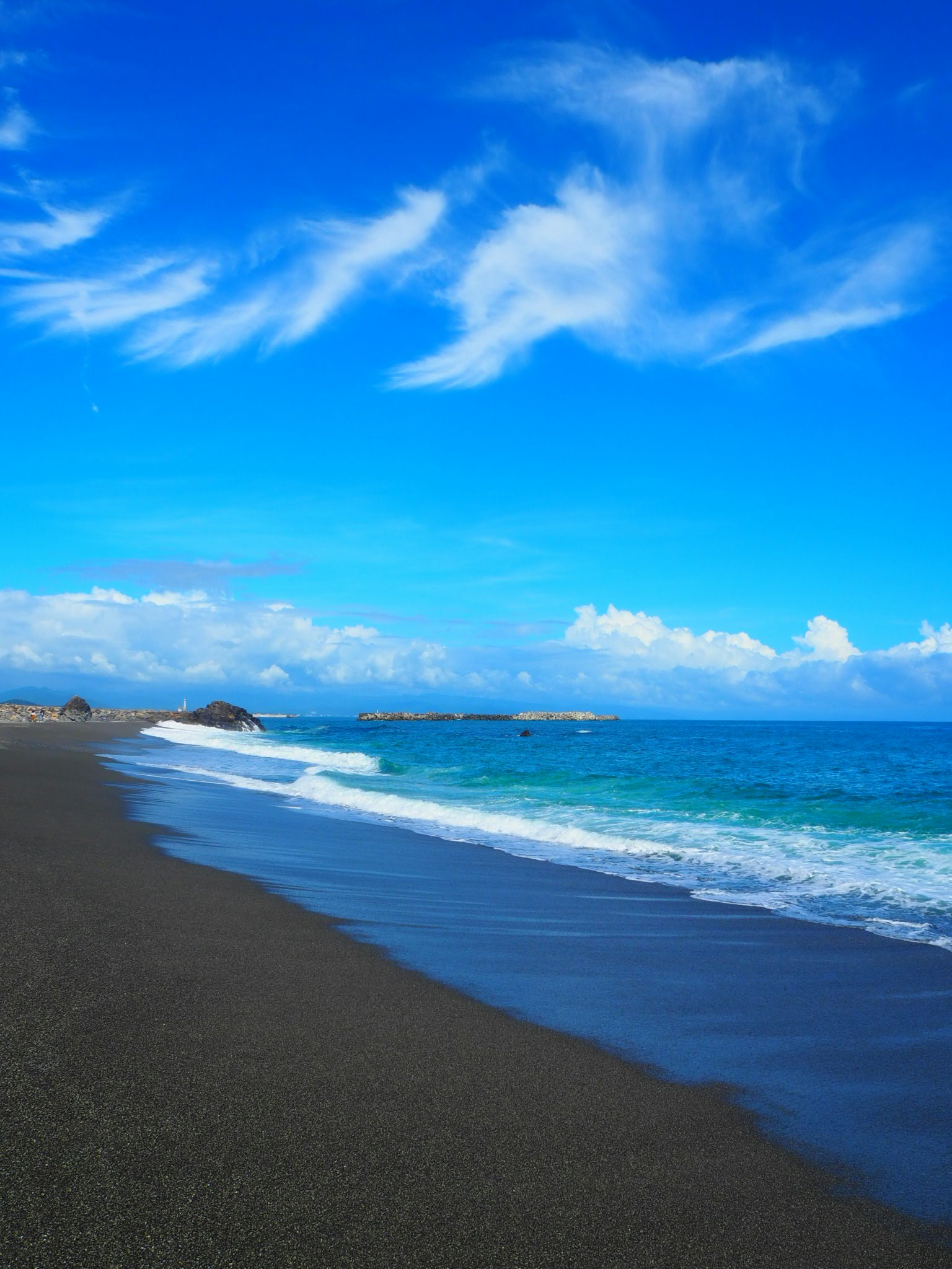 ชายหาดทรายดำกับมหาสมุทรสีน้ำเงินใต้ท้องฟ้าสว่างและเมฆขาว