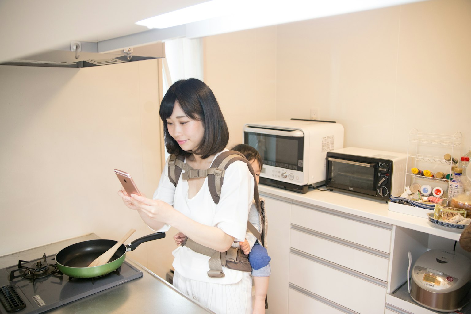 Mujer cocinando en la cocina mientras usa un teléfono inteligente con un niño en su espalda
