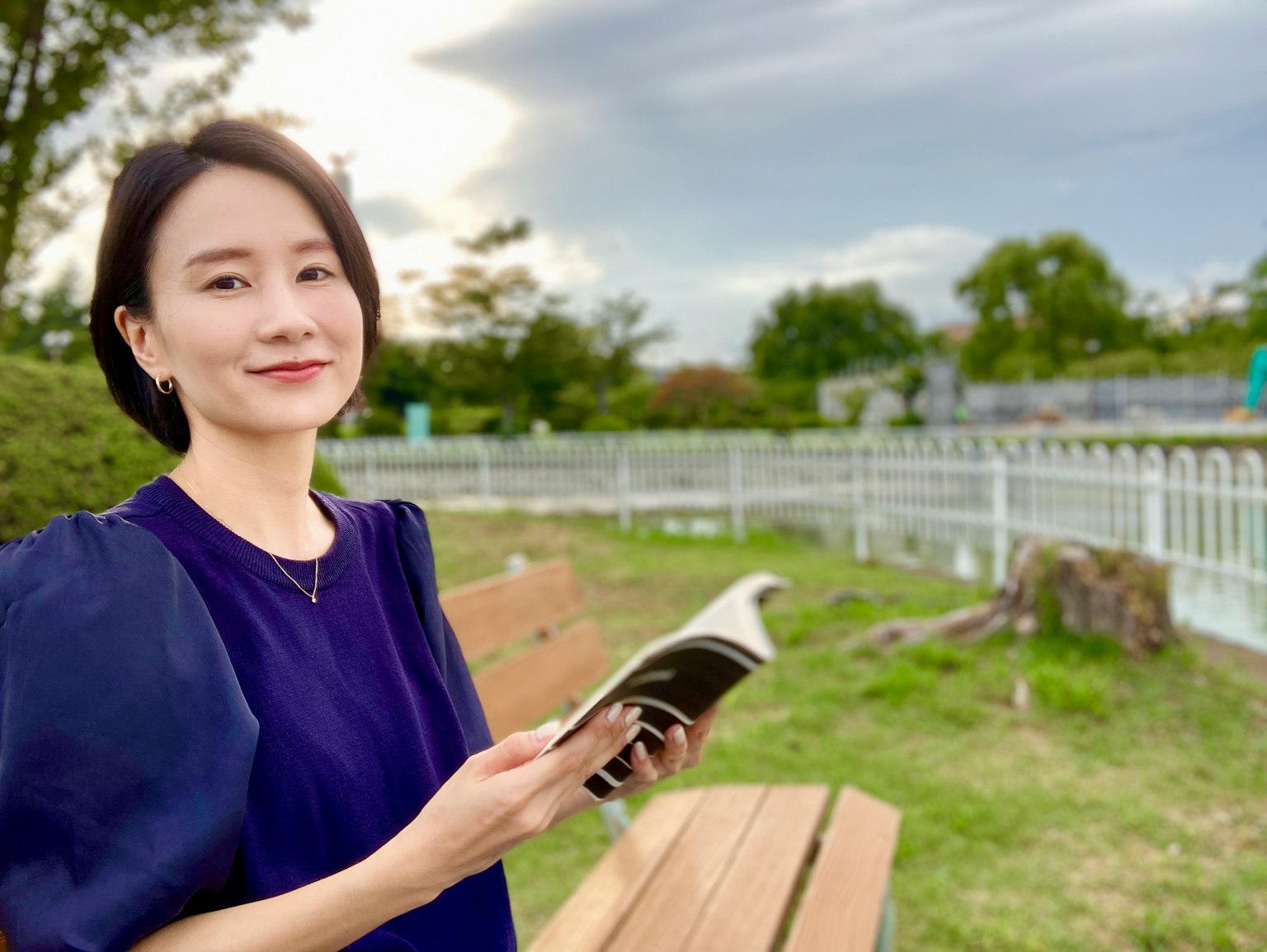 Wanita tersenyum memegang buku di taman dengan langit biru dan pemandangan hijau