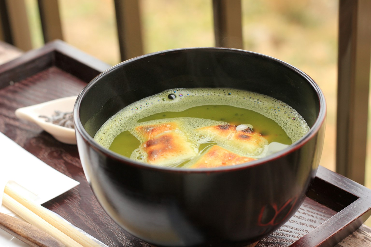Schwarze Schüssel mit grünem Teesuppe und Essstäbchen auf einem Holztablett