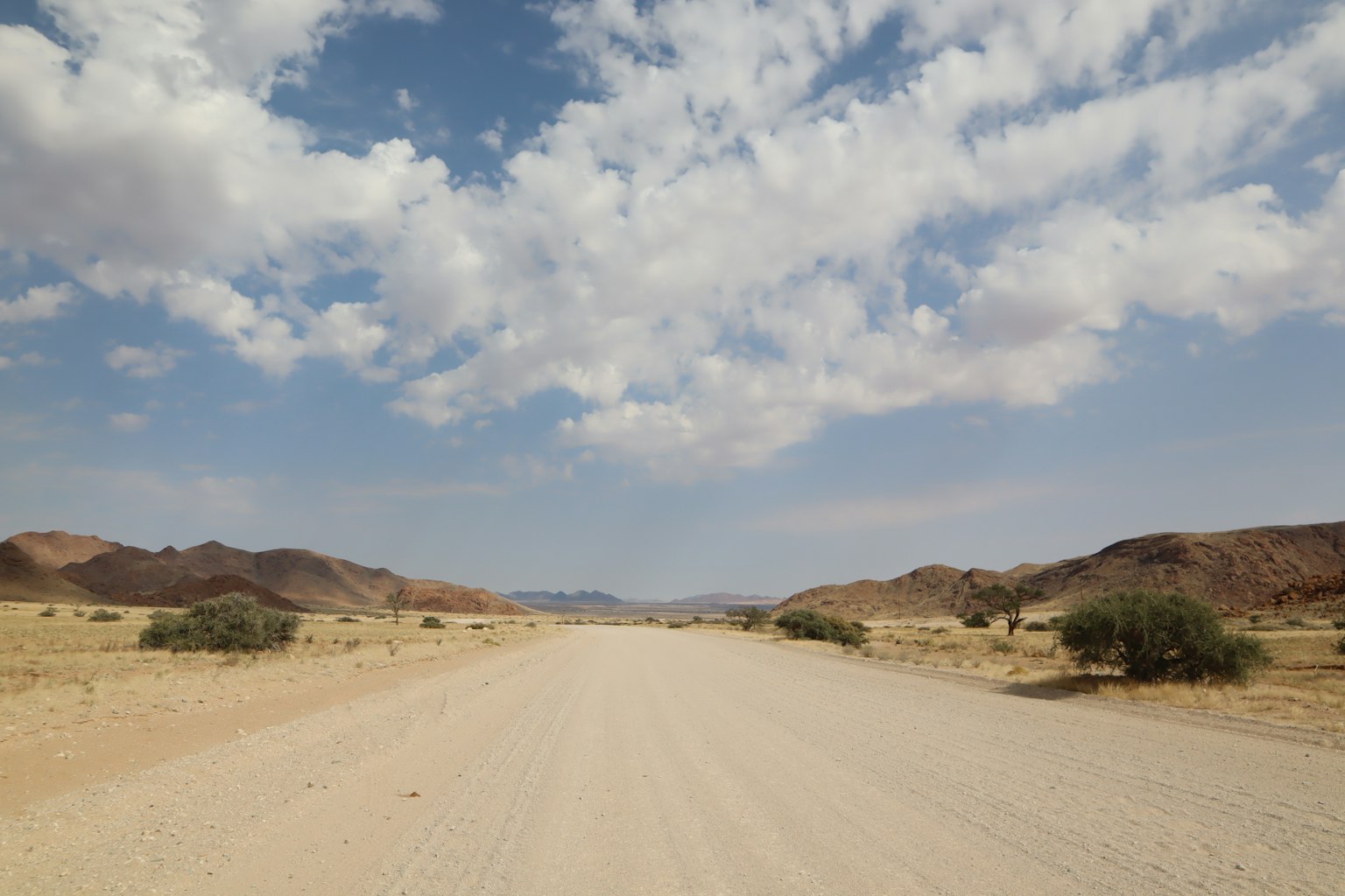 Strada sterrata che si estende attraverso un paesaggio arido con cielo azzurro e nuvole
