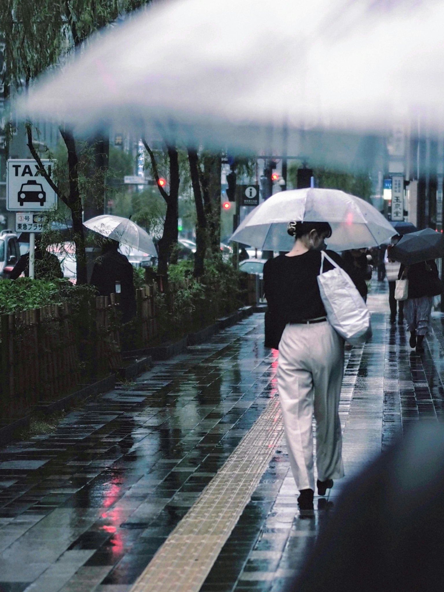 Femme marchant sous la pluie avec un parapluie blanc portant une tenue noire sur un pavé réfléchissant dans un cadre urbain