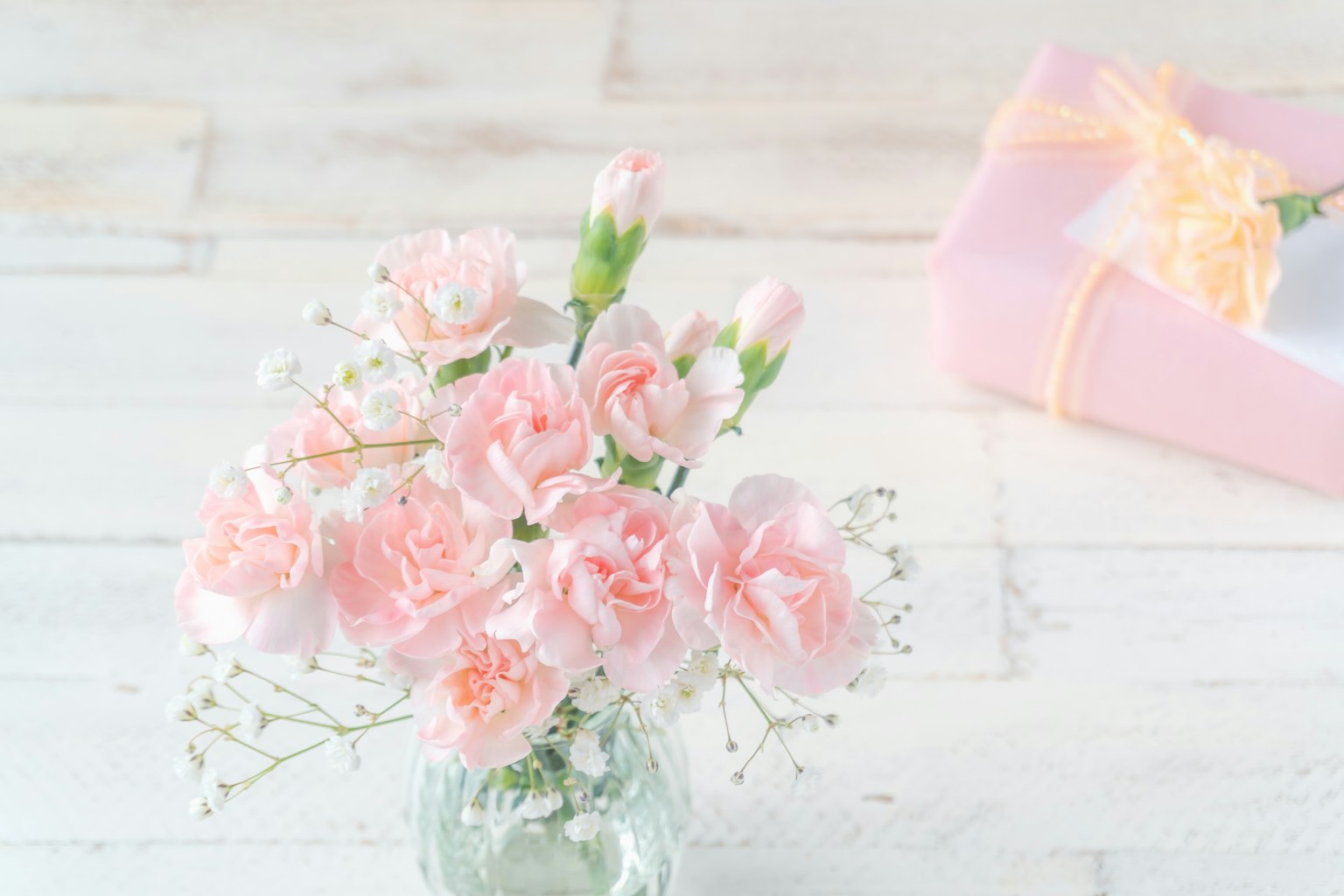 Un bouquet de œillets roses doux et de petites fleurs blanches dans un vase en verre avec une boîte-cadeau rose en arrière-plan
