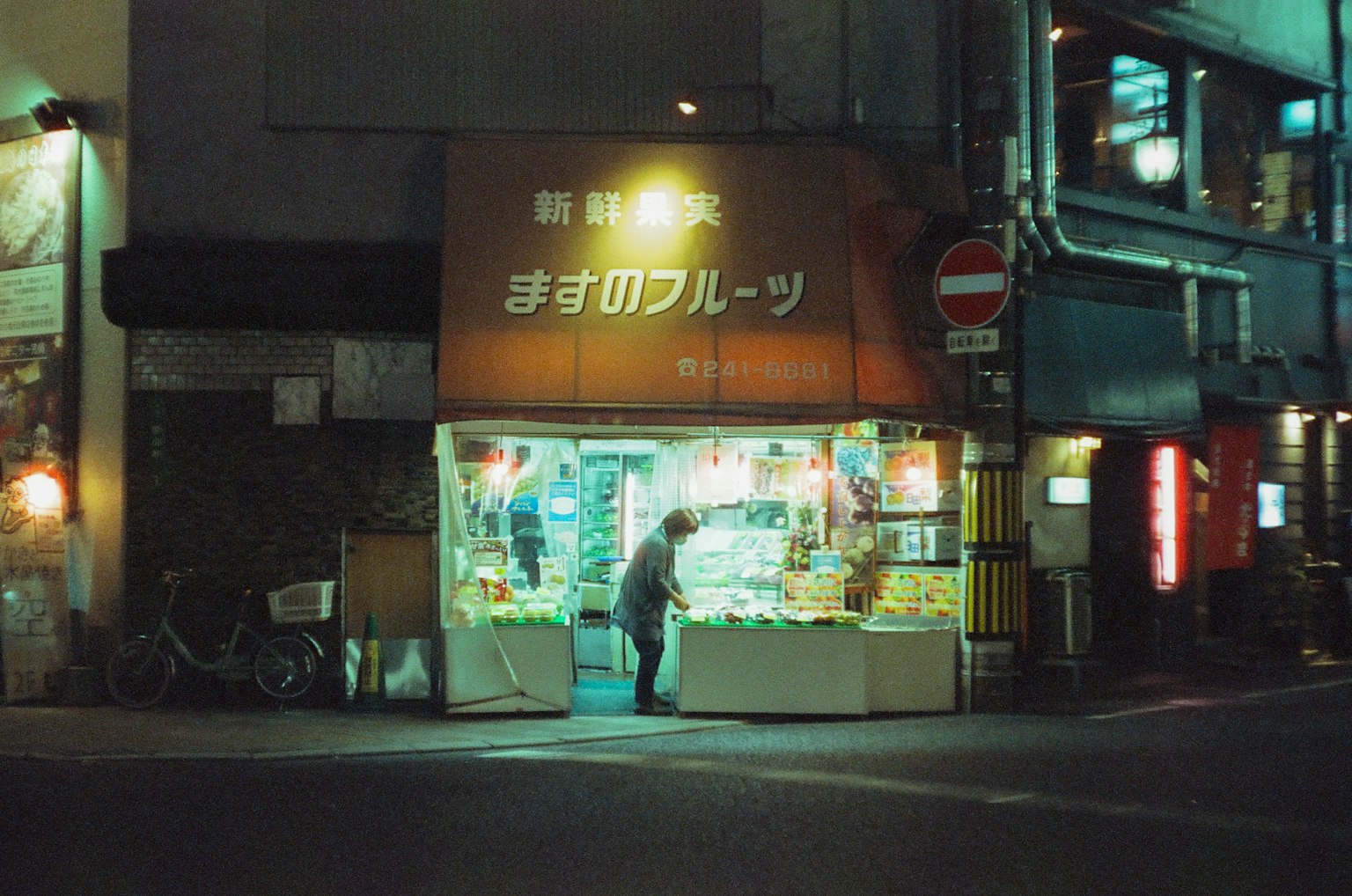 Exterior de una tienda de frutas en una esquina de la calle por la noche con un vendedor