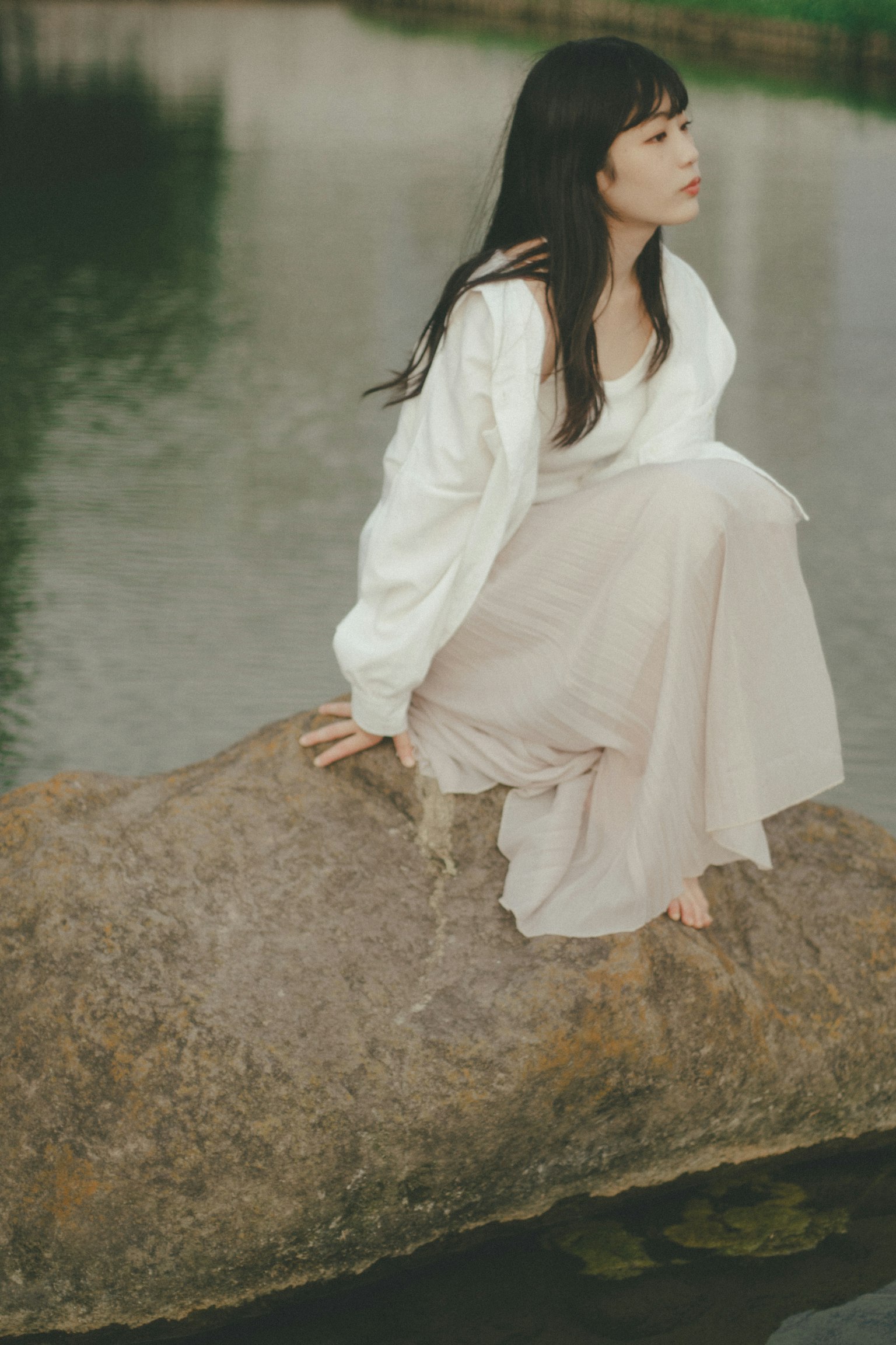 หญิงสาวนั่งอยู่บนก้อนหินข้างน้ำสวมเสื้อสีขาวและกระโปรงสีอ่อนในบรรยากาศธรรมชาติที่เงียบสงบ