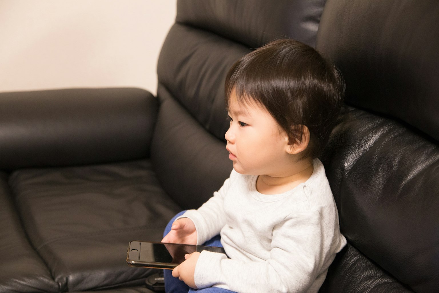 Un niño pequeño sentado en un sofá negro sosteniendo un teléfono inteligente