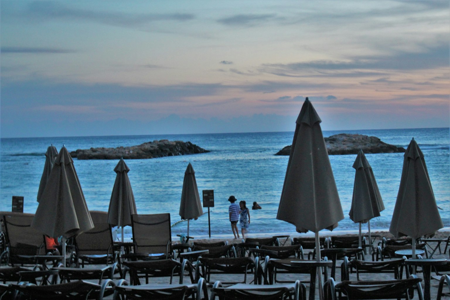 Scena di spiaggia con ombrelloni e sedie che si affacciano sul mare e sulle rocce al tramonto
